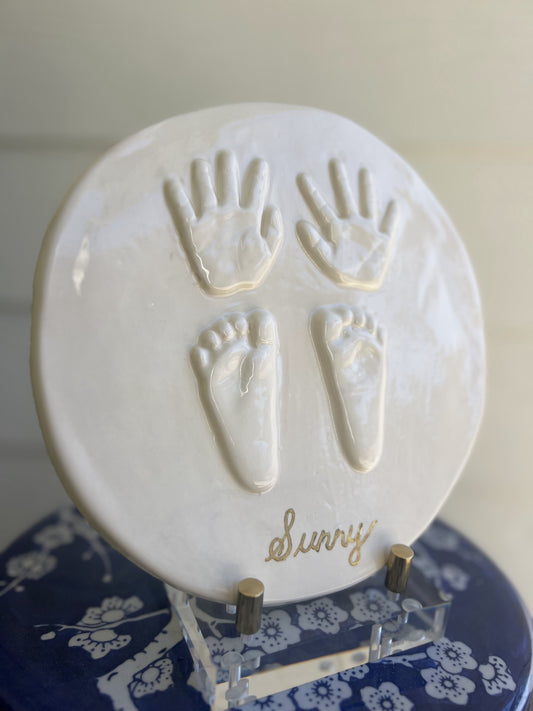 Hand-cast three-dimensional ceramic round plaque featuring a child’s handprints and footprints, glazed and kiln fired with personalized name.